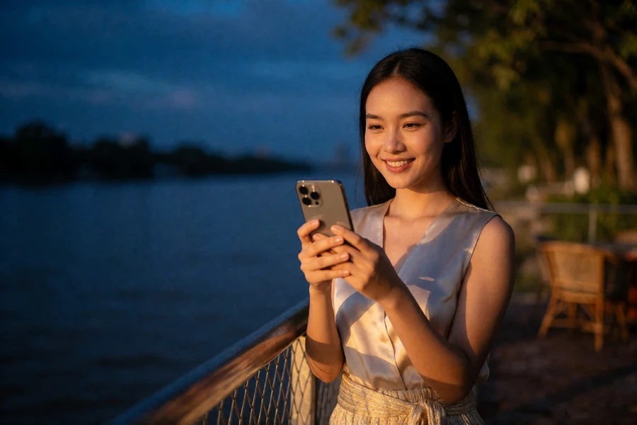 A Thai woman smiling at her phone during a video call on a riverside terrace at dusk, illustrating live video connection across a language gap